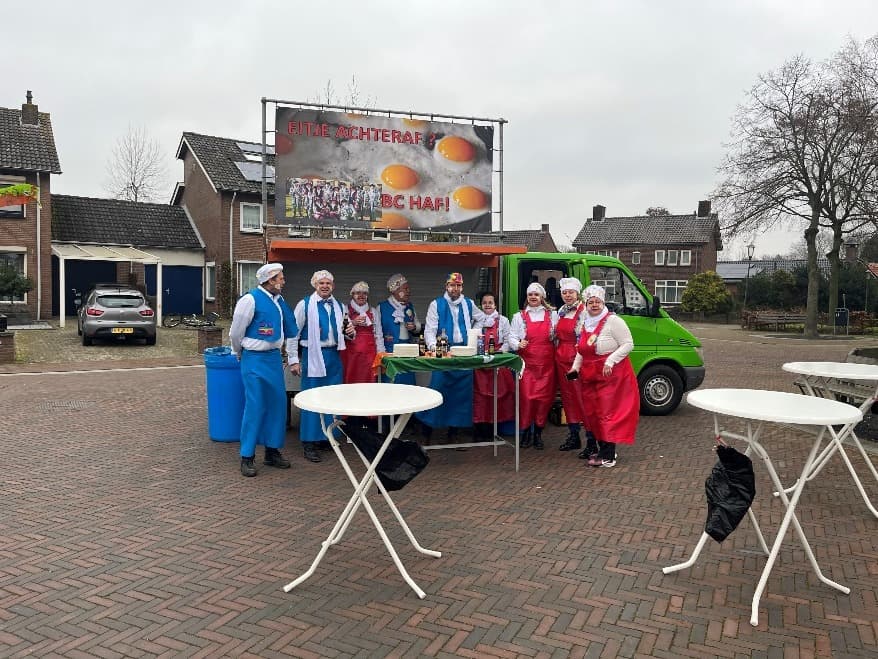 Carnavalsgroep in kostuums bij een busje met een bord