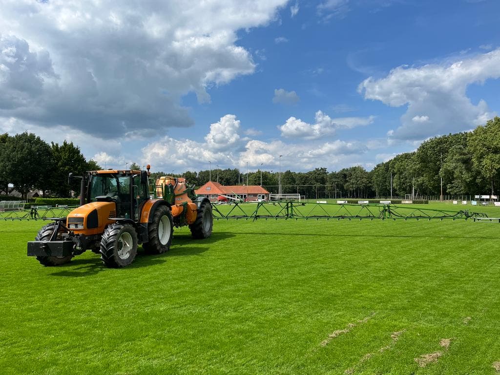 Oranje trekker met veldspuit op het voetbalveld, clubhuis op de achtergrond